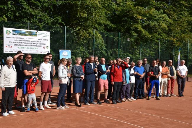 Grupa ludzi - kobiet i mężczyzn - stoi na korcie tenisowym i patrzy w jednym kierunku.