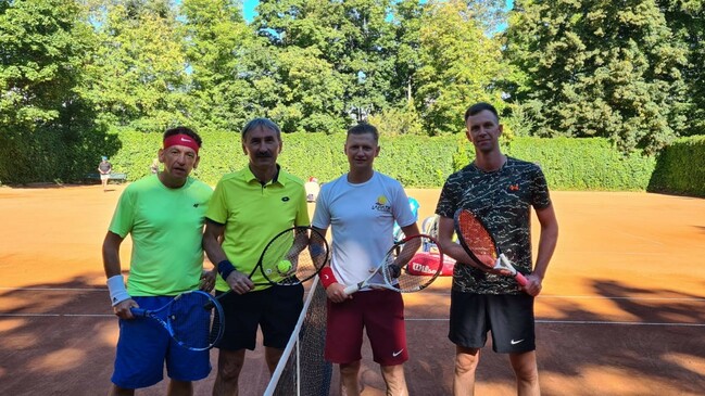 czterech mężczyzn z rakietami tenisowymi stoi na korcie tenisowym. W rękach trzymają rakiety tenisowe, mają na sobie sportowe stroje. Patrzą w obiektyw aparatu i się uśmiechają.